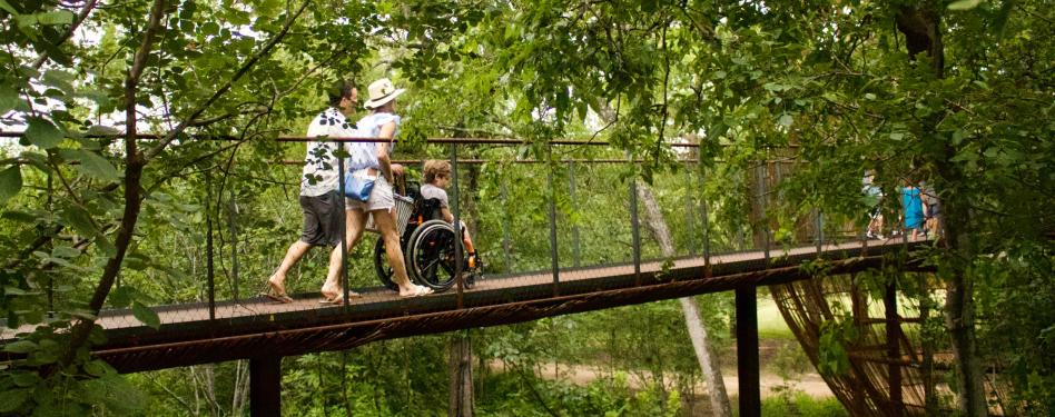 Two parents roll their child's wheelchair along a footbridge in a forested area.