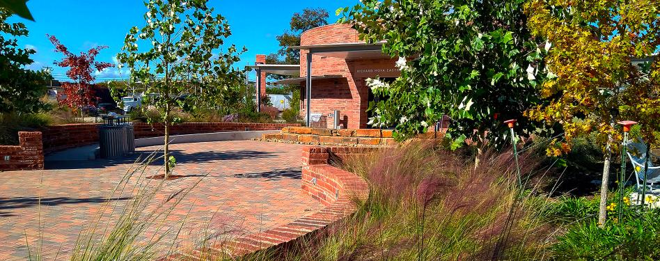 Red brick walkway leads to low circular building, with trees and grasses throughout the area.