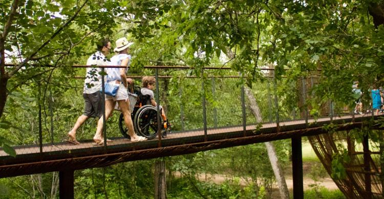 Two parents roll their child's wheelchair along a footbridge in a forested area.